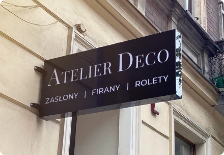 Szyld zewnętrzny – Po prawej stronie widać elegancki, czarny szyld „Atelier Deco” nad wejściem do sklepu, podkreślający luksusowy charakter marki zajmującej się dekoracją wnętrz.