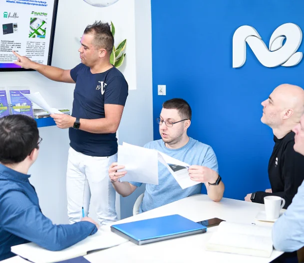 Na zdjęciu widzimy spotkanie zespołu, w którym biorą udział cztery osoby. Jedna z nich stoi przy ekranie, wskazując na prezentowaną treść, podczas gdy pozostałe trzy osoby siedzą przy stole, trzymając w rękach dokumenty