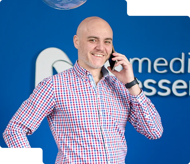 Zdjęcie przedstawia uśmiechniętego mężczyznę prowadzącego rozmowę telefoniczną. Ubrany jest w koszulę w kratę i zegarek na lewym nadgarstku. W tle widać niebieską ścianę z logo Media Essence, 