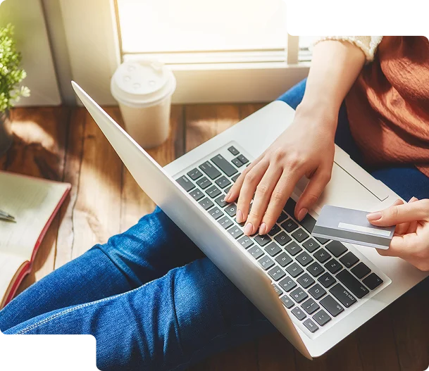 Na zdjęciu widoczna jest osoba siedząca na podłodze, używająca laptopa. Trzyma w ręce kartę kredytową i zapewne dokonuje zakupu online. Obok znajduje się kubek z napojem